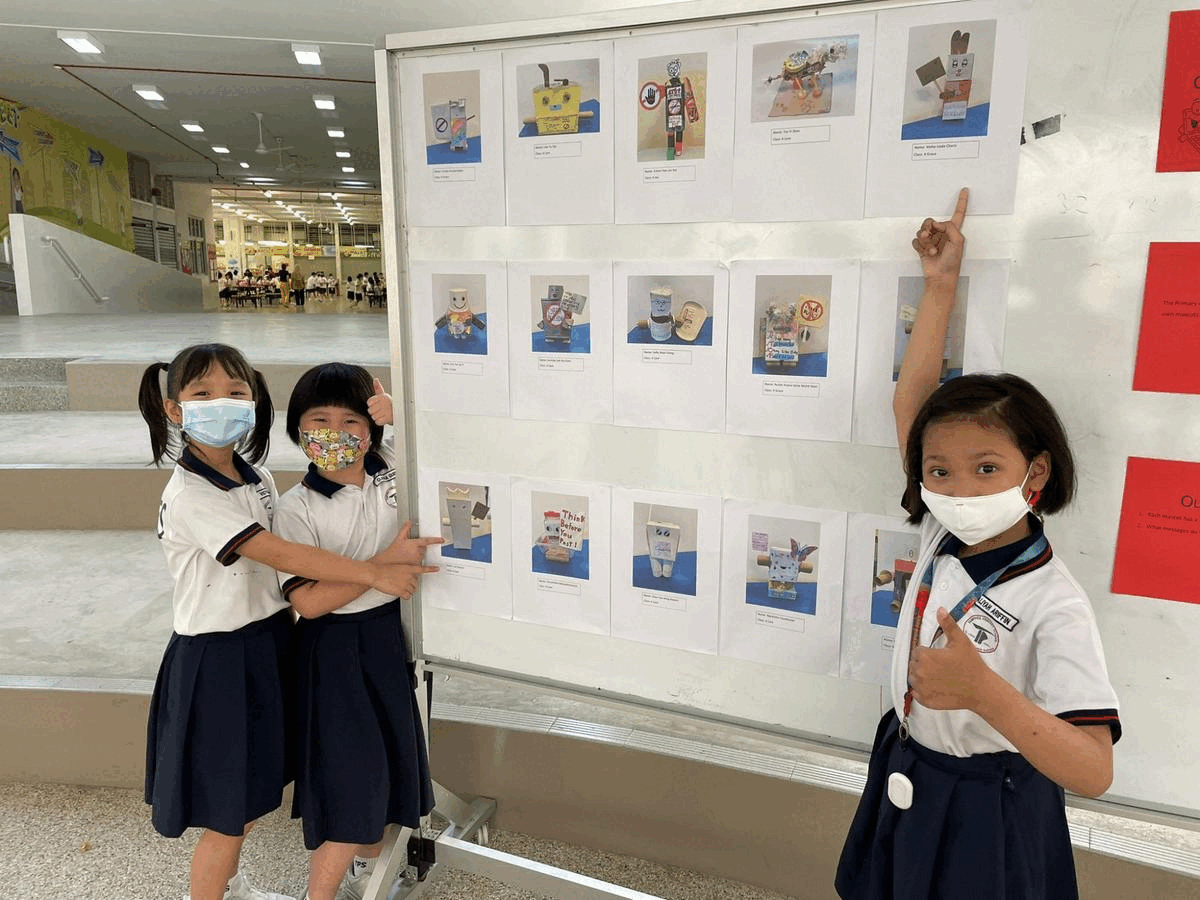 Three young girls in school uniforms and face masks pose proudly beside a display board of student craft photos.